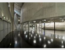 Großer, moderner Innenraum mit Betonwänden und hochglänzendem dunklem Boden. Es gibt breite Treppen, Rundsäulen und eine kurvige Wand, begleitet von eingebauten Deckenleuchten für gleichmäßige Beleuchtung.