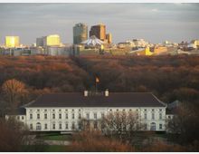Luftaufnahme eines weißen, historischen Gebäudes mit großen Fenstern und braunem Dach, umgeben von Bäumen im Tiergarten in Berlin. Im Hintergrund sind moderne Hochhäuser sowie das Dach des Sony Centers erkennbar.