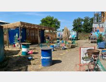 Ein Außenbereich mit rustikalem Ambiente, bestehend aus Holzhütten, Metallfässern als Tische, verschiedenen Gartenstühlen und Dekorationen wie pinken Flamingofiguren. Im Hintergrund stehen bunt bemalte Container, umgeben von Bäumen und einem klaren Himmel.