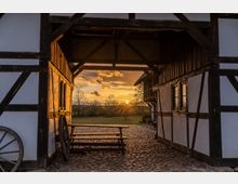 Blick durch den Eingang eines historischen Fachwerkhofs, dessen schwarzes Holzgebälk von weiß verputzten Wänden eingerahmt wird, auf eine Wiese im Sonnenuntergang. Der gepflasterte Innenhof ist mit einer Holztischgruppe, einem Wagenrad und umgebenden Bäumen gestaltet.