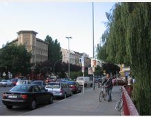 Straßenszene in einer Stadt mit mehrspuriger Straße und parkenden Autos, flankiert von historischen Wohngebäuden mit aufwendigen Fassaden. Rechts ein bepflanzter Gehweg mit Bäumen, darunter eine Trauerweide, sowie ein Mann mit einem Fahrrad.