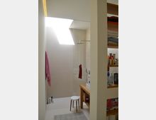 Helles, modernes Badezimmer mit einer begehbaren Dusche, einer Glasabtrennung und einem Oberlicht, das viel Tageslicht hereinlässt. Rechts stehen Holzregale mit Pflegeprodukten, daneben ein Waschtisch mit einer Keramikwaschschüssel und ein kleiner Hocker.