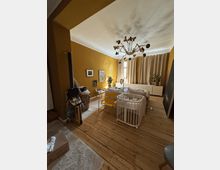 Cozy bedroom with mustard-yellow walls, wide wooden plank flooring, and a large mid-century style chandelier on the ceiling. A bed sits in front of a curtained window beside a long white cabinet with potted plants, with a crib and chairs in the foreground.