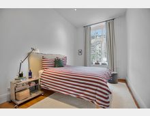 Schlafzimmer mit weiß gestrichenen Wänden, großem Bett mit weiß-rot gestreifter Bettwäsche und einem Nachttisch mit Leselampe, Büchern und kleinen Dekorationen. Ein großes Fenster mit Vorhängen lässt Tageslicht herein und bietet Blick auf Bäume und Gebäude im Hintergrund.