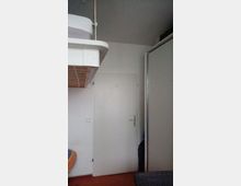 Kleines Zimmer mit weiß gestrichenen Wänden, einer weißen Tür in der Mitte und einem hohen Hochbett in der oberen linken Ecke. Rechts befindet sich ein großer, weißer Kleiderschrank mit einer Schiebetür.
