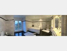 Geräumiges Badezimmer mit weiß gefliesten Wänden und dunklem glänzendem Boden. In der Mitte ein großes, eingelassenes Eckbad mit Stufen, daneben eine runde Duschkabine. Links ist ein großes Fenster, rechts ein breiter Waschtisch mit Spiegeln und Ablagen.