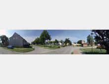 Straßenkreuzung in einem Wohngebiet mit Kopfsteinpflasterstraßen, umgeben von Einfamilienhäusern, Bäumen und Grünflächen. Im Hintergrund sind gelb und weiß gestrichene Gebäude zu sehen, während Verkehrsschilder und ein parkendes Auto rechts im Bild sichtbar sind.