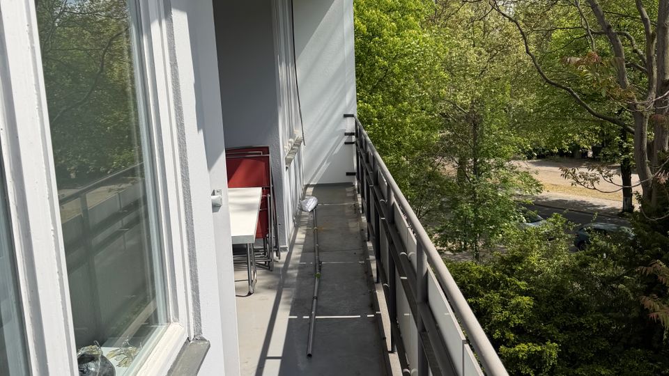 Schmaler Balkon an einem Wohngebäude mit grauem Boden, Metallgeländer und großer Fensterfront; am Ende stehen ein kleiner Tisch und Stühle. Der Balkon blickt auf dichtes Grün mit hohen Bäumen und eine Straße im Hintergrund.