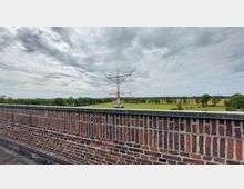 Backsteinmauer im Vordergrund, dahinter eine große rote und weiße Antennenstruktur in einer grünen Wiesenlandschaft mit Bäumen am Horizont unter teilweise bewölktem Himmel.