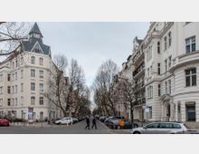 Straßenszene in einem Wohnviertel mit klassischen Altbauten, verziert mit Balkonen und Erkern. Die Straße ist von kahlen Bäumen gesäumt, mit geparkten Autos auf beiden Seiten und zwei Fußgängern, die die Straße überqueren.