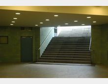 Treppe in einem unterirdischen Durchgang mit grünen gefliesten Wänden, blauer Tür und Deckenbeleuchtung aus eingelassenen, runden Lampen. Die Treppe führt zu einem helleren Außenausgang.