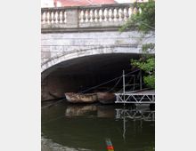 Steinbrücke mit Balustrade über einem Wasserlauf, darunter zwei rostige Boote und eine kleine Metallplattform am Ufer. Im Vordergrund reflektiert das Wasser, während Äste eines Baumes von rechts ins Bild ragen.