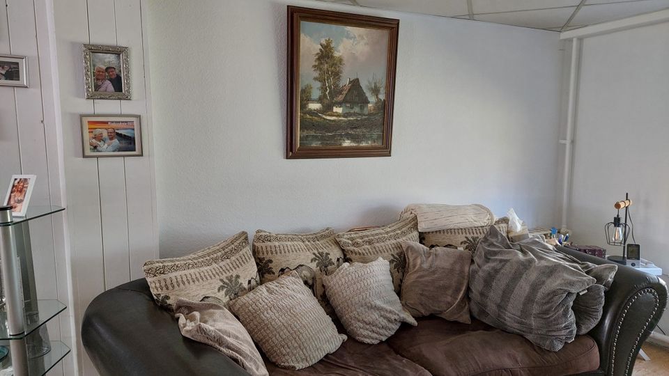 Wohnzimmerbereich mit dunklem Sofa, vielen Kissen und Decken vor einer weißen Wand. Darüber hängen ein großes gerahmtes Landschaftsbild und mehrere kleinere gerahmte Fotos; links steht ein Glasregal, rechts ein kleines Beistelltischchen mit Lampe.