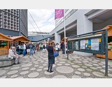 Ein Platz in einer Stadt mit zahlreichen runden Bodenplatten, umgeben von modernen Gebäuden. Es sind mehrere Holzstände wie auf einem Markt sowie Menschen in unterschiedlicher Bewegung zu sehen; rechts befindet sich eine Infotafel.
