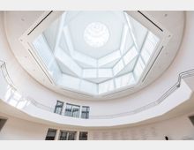 Blick auf das Innere eines modernen Museums mit einem großen, achteckigen Glasdach, das viel Tageslicht hereinlässt. Die Wände sind weiß und mit einer umlaufenden Galerie versehen, während im unteren Bereich Schrifttafeln sichtbar sind.