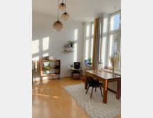 Helles Wohnzimmer mit großen Fenstern, durch die Licht einfällt, einem Holzboden und einer minimalistischen Einrichtung. Im Raum befinden sich ein Holztisch mit Stühlen auf einem gemusterten Teppich, ein Regal mit Büchern und Dekoration sowie Zimmerpflanzen und eine moderne Hängelampe mit drei kugelförmigen Leuchten.