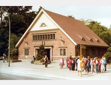 Backstein-Gebäude mit Satteldach, Holzelementen im Giebel und mehreren Fenstern, beschriftet mit "Bhf. Oskar-Helene-Heim". Vor dem Gebäude befinden sich eine Gruppe von Menschen sowie ein Gehweg, umgeben von Bäumen im Hintergrund.