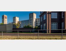 Industriekomplex mit markanter Backsteinarchitektur im Vordergrund und einem großen Gebäude mit zwei Uhrtürmen im Hintergrund, umgeben von Bäumen und einer Mauer. Im Vordergrund verlaufen eine Straße und ein Geländer.