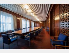 Langer Konferenzraum mit hölzerner Wandverkleidung, einem ovalen Tisch in der Mitte und blauen Polsterstühlen. Die Decke ist mit einem Muster aus runden Elementen gestaltet, und große Fenster mit Spitzenvorhängen lassen Tageslicht herein.