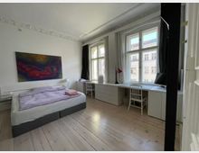 Helles Schlafzimmer mit Holzdielenboden, einem Doppelbett mit heller Bettwäsche und einem großen farbigen Gemälde an der weißen Wand. Vor den großen Fenstern steht ein langer weißer Schreibtisch mit zwei Stühlen und einer roten Leselampe.
