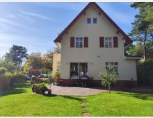 Zweigeschossiges Einfamilienhaus mit cremefarbener Fassade, roten Fensterläden und steilem Satteldach. Im Vordergrund ein großer Garten mit Rasenfläche, Zierpflanzen in Töpfen und einer Terrasse aus Steinplatten; im Hintergrund Bäume und vereinzelte Waldflächen.