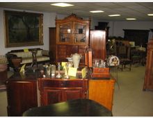 Ein Raum mit antiken Möbeln, darunter Vitrinen, Kommoden, Polstermöbel und dekorative Objekte wie Vasen und Kerzenhalter. An der Wand hängt ein großformatiges Gemälde in einem goldenen Rahmen.