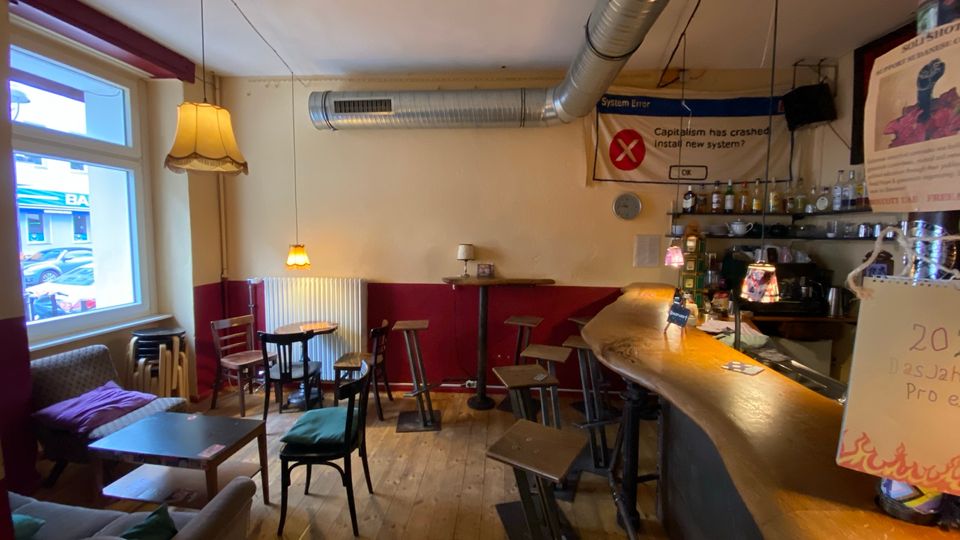 Gemütlicher Bar- und Caféinnenraum mit Holzfußboden, kleiner Sitzecke am Fenster, gemischten Tischen und Hockern sowie einer geschwungenen Theke. Sichtbare Lüftungsrohre an der Decke, Hängelampen und Regale mit Flaschen hinter der Bar prägen das Interieur.