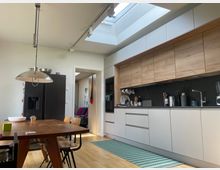 Moderne Wohnküche mit langen weißen und holzfarbenen Einbauschränken, dunkler Arbeitsfläche und Spüle sowie schwarzem Kühlschrank neben einem Esstisch aus Holz. Ein großes Oberlicht im Dach und Schienenstrahler sorgen für viel Tageslicht, im Hintergrund führt eine Türöffnung in einen angrenzenden Raum.