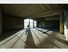 Leerer, rohbauartiger Raum mit sichtbarer Betonstruktur und großem Fenster, durch das Sonnenlicht hereinströmt. Im Hintergrund steht eine Person vor den Fenstern, wodurch lange, scharfe Schatten auf den Boden fallen.