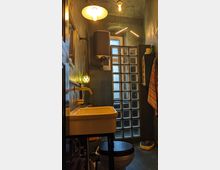 Kleines Badezimmer mit dunklen Wänden, einem Waschbecken mit goldfarbener Armatur, einer schwarzen Toilette und einer Wand aus Glasbausteinen. Die Beleuchtung besteht aus hängenden, warmweißen Lampen sowie einer Kugellampe an der Wand.