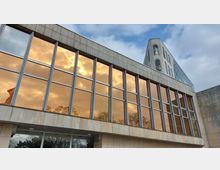 Ein modernes Gebäude mit großen, rechteckigen Glasfenstern, die den orangefarbenen Himmel bei Sonnenuntergang reflektieren. Im Hintergrund ist ein Teil eines Gebäudeteils mit dekorativen Figuren und einer gläsernen Dachstruktur sichtbar.