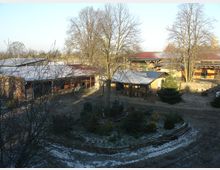Ein Bauernhof mit mehreren Stallgebäuden und offenen Unterständen, umgeben von kahlen Bäumen im Winter. Der Boden ist teilweise mit Schnee bedeckt, und ein Gartenbereich liegt in der Mitte des Geländes.