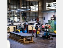 Innenraum einer Werkstatt mit industriellem Charakter. Zu sehen sind Arbeitsflächen, Werkzeuge, ein Schraubstock, eine Werkbank, ein Kompressor, und eine Metallplatte; im Hintergrund befinden sich große Fenster und Regale mit Arbeitsmaterialien.