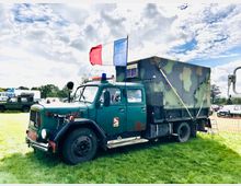 Ein grüner, militärisch anmutender Lastwagen mit Tarnmuster auf dem Aufbau und einer blauen und roten Sirene auf dem Dach steht auf einer Wiese. Auf dem Dach flattert eine französische Flagge, im Hintergrund sind weitere Fahrzeuge und ein wolkiger Himmel zu sehen.