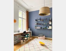 Bright study room with a tall arched window, ornate white ceiling molding, and a blue accent wall with long shelves displaying toys and books. A long desk with chairs sits by the window, and a light rug with a small low table and floor cushion covers the wooden floor.