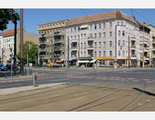 Straßenkreuzung in einer städtischen Umgebung mit Straßenbahnschienen und mehrstöckigen Wohnhäusern im Hintergrund. Die Gebäude verfügen über Balkone, Geschäfte im Erdgeschoss und eine beige Fassade; darunter sind ein Fotoladen und ein Restaurant zu erkennen.