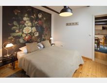 Schlafzimmer mit einem Doppelbett, beige Bettwäsche und Nachttischen mit Lampen, vor einer großen Wand mit floralem Kunstmotiv. Im Hintergrund ist eine Tür zu einer Küche mit Esstisch und Stühlen sichtbar.