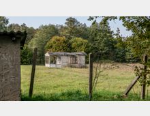 Eine verlassene Holzhütte mit zerbrochenen Fenstern steht auf einer Wiese vor einem Hintergrund aus dichten Bäumen. Im Vordergrund befinden sich Zaunpfähle und ein verputztes Gebäude mit Wellblechdach am linken Bildrand.