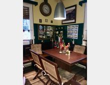 Gemütlicher Essbereich mit holzvertäfeltem Tisch, Korbstühlen und Bank mit Fellkissen. An den Wänden hängen eine Uhr, mehrere Dekorationsbilder und Spiegel, darunter Vasen mit Kunstblumen, sowie ein Wandregal mit silbernen Behältern.