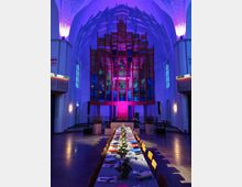 Innenraum einer modern gestalteten Kirche mit hoher Gewölbedecke, farbiger Beleuchtung in Lila- und Blautönen und einem auffälligen, abstrakten roten Kunstwerk am Altarbereich. Mittig steht eine lange, festlich gedeckte Tafel mit Kerzen und Blumenschmuck.
