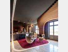 Moderner Lounge-Bereich mit geschwungenen Holzpaneelen an einer Wand, einer großen Fensterfront, hellen Polstersesseln und einem runden Tisch auf einem roten Teppich. Beleuchtungselemente an der Decke und um den Teppichrahmen sowie eine Bar mit Barhockern im Hintergrund.