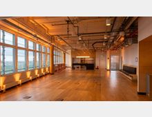 Spacious modern loft-style office or event space with a wide wooden floor, exposed ceiling pipes and suspended lights, and floor-to-ceiling windows along one wall overlooking a cityscape. A small kitchenette and high-top seating area sit at the back, with built-in bench seating along the opposite wall.