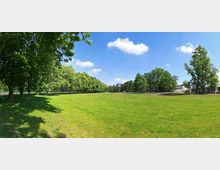 Weitläufiger Park mit einer großen, gepflegten Grünfläche, umgeben von zahlreichen Bäumen. Im Hintergrund sind Gebäudestrukturen und ein blauer Himmel mit vereinzelten Wolken sichtbar.