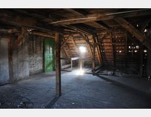 Ein leerer Dachboden mit Holzträgern und einem schrägen Ziegeldach, durch das leichtes Tageslicht einfällt. An der Rückwand befindet sich eine gemauerte Wand mit einer grünen Tür, der Boden ist aus Holzplanken.