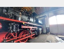 Dampf-Lokomotive in einer Werkstatthalle, mit roten Rädern und einem schwarzen Kessel, umgeben von Backsteinwänden und einer geöffneten großen Rolltür. Das Dach zeigt eine Holzbalkenkonstruktion.