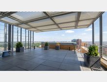 Rooftop observation terrace with large gray floor tiles, metal railings, and a slatted canopy overhead, lined with square planters. A panoramic city skyline and a large green park are visible beyond the glass and open edge.