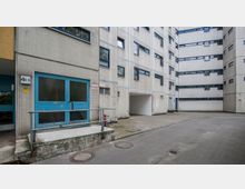 Mehrstöckiges Wohngebäude aus Beton mit kleinen Fenstern und einer blau gestrichenen Eingangstür. Im Vordergrund ein gepflasterter Hof mit Entwässerungsschächten und ein Zugang mit Rampe.