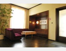 Wohnbereich mit einer dunklen Ecksofagarnitur, einem kleinen Tisch mit Tischdecke und einer großen Topfpflanze links. Ein großes Fenster mit hellen Vorhängen lässt Tageslicht herein; die Wandverkleidung aus Holz enthält einen dekorativen Wandspiegel und eine Lampe.