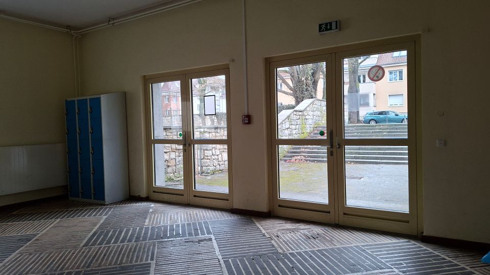 Innenraum eines Gebäudeeingangs mit hellem Anstrich, blauem Spindschrank und großflächigem Fliesenmusterboden. Zwei Doppeltüren aus Glas führen nach draußen zu einer Treppe neben einer Steinmauer und Blick auf Wohnhäuser und parkende Autos.