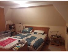 Ein Schlafzimmer mit einem Doppelbett aus Holz, ausgestattet mit blauer Bettwäsche und mehreren Kissen. Im Raum befinden sich außerdem ein Nachttisch mit Lampe, ein Bücherregal, sowie eine Wickelkommode mit Babyutensilien, und die Schrägen der Dachgeschossdecke sind sichtbar.